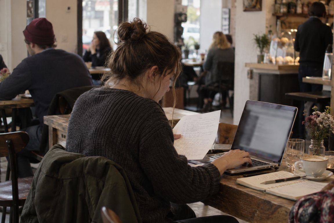 Frau liest in einem Café handschriftliche Notizen und tippt dabei auf ihrem Laptop.
